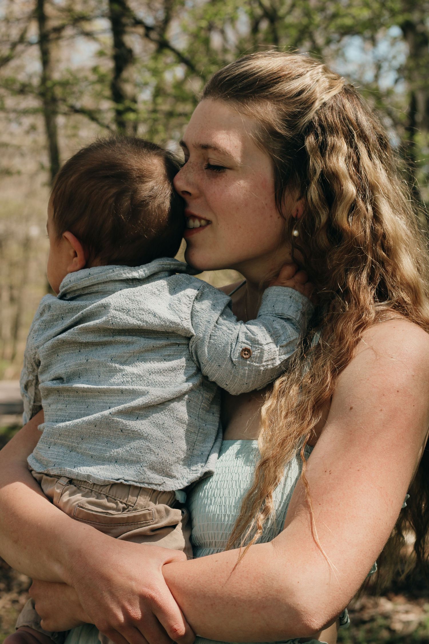 A young woman with curly hair tenderly holds and smiles at a baby dressed in light blue outdoors.