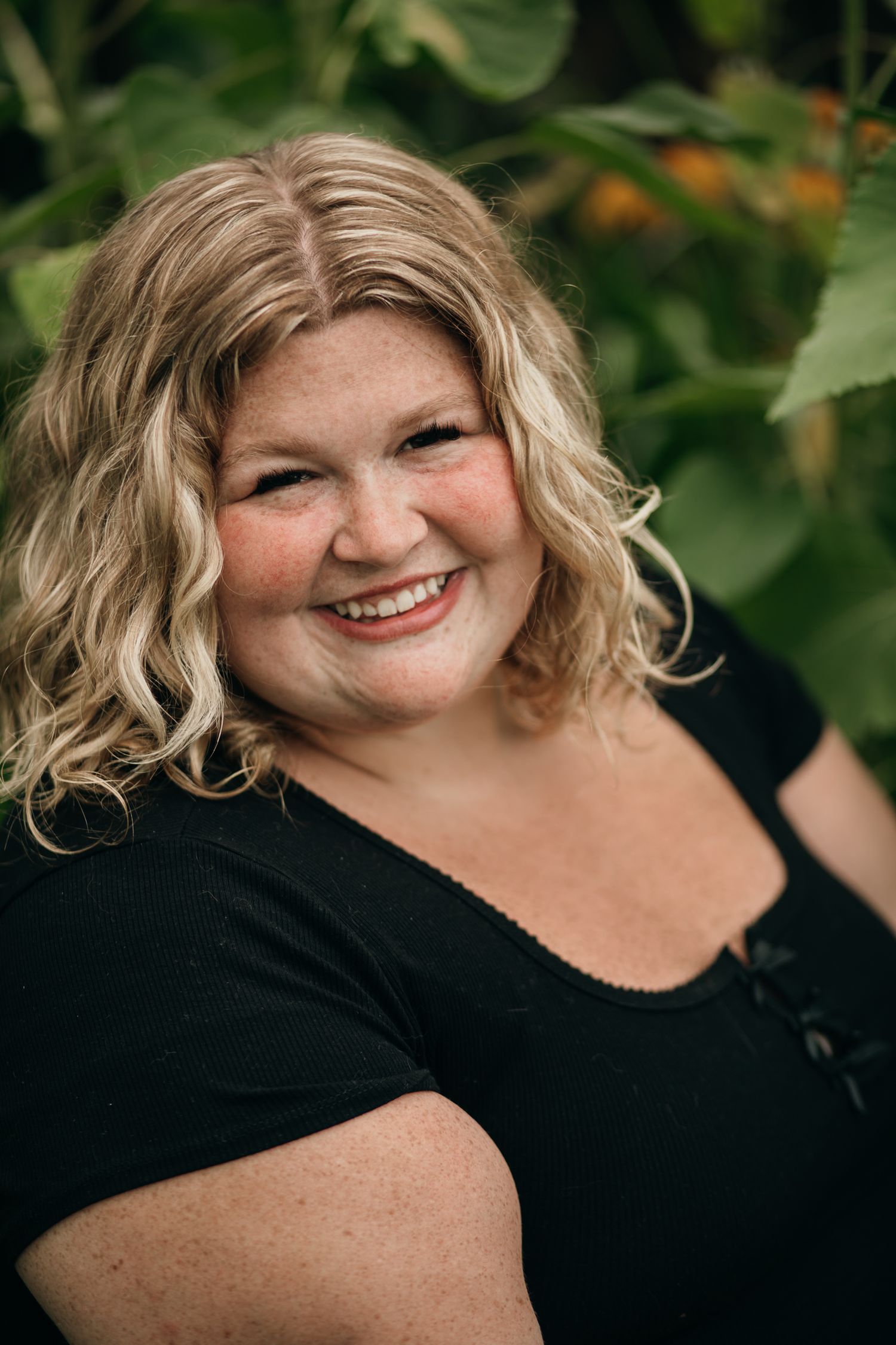 Smiling blonde woman with freckles wearing a black top, posing outdoors among green sunflower leaves.