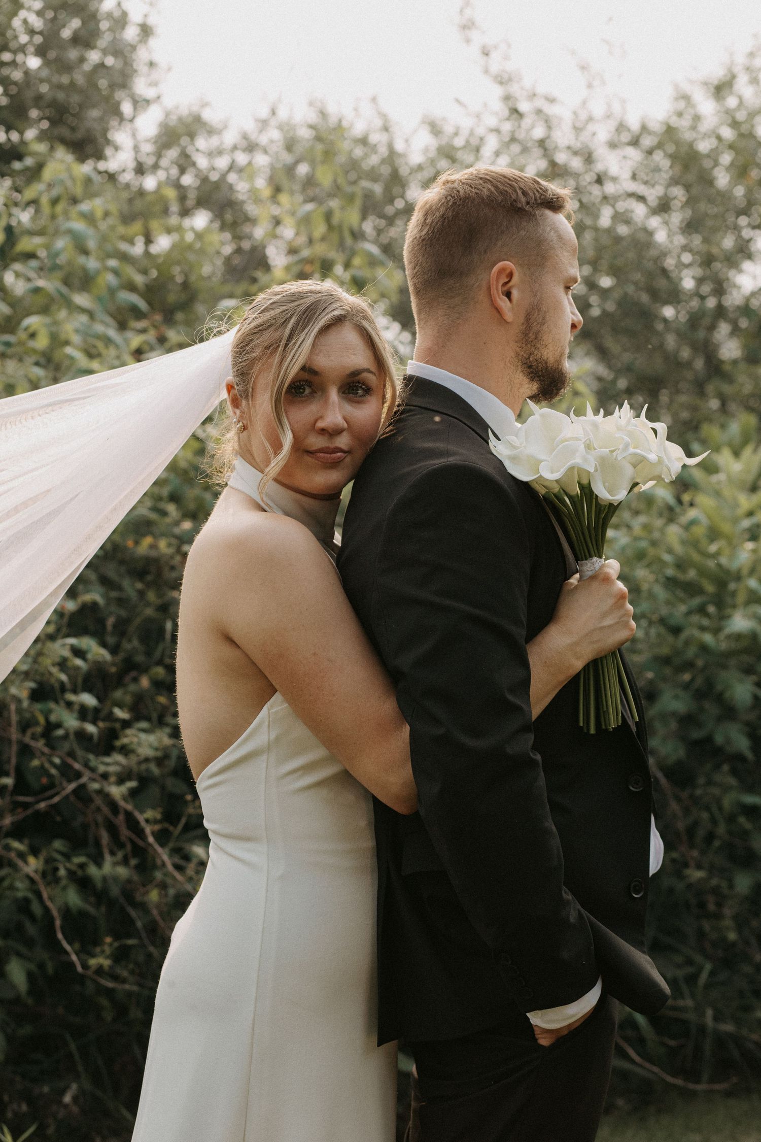 Bride in sleek white gown hugs groom from behind as veil flows, groom holds white bouquet.