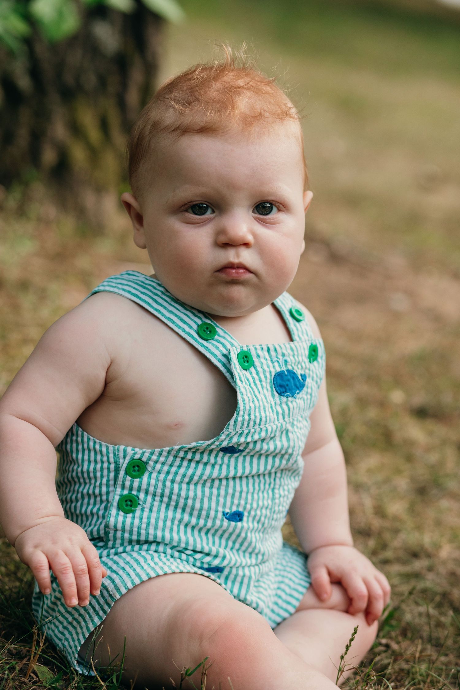 Adorable red-haired baby in striped green romper sitting on grass outdoors.