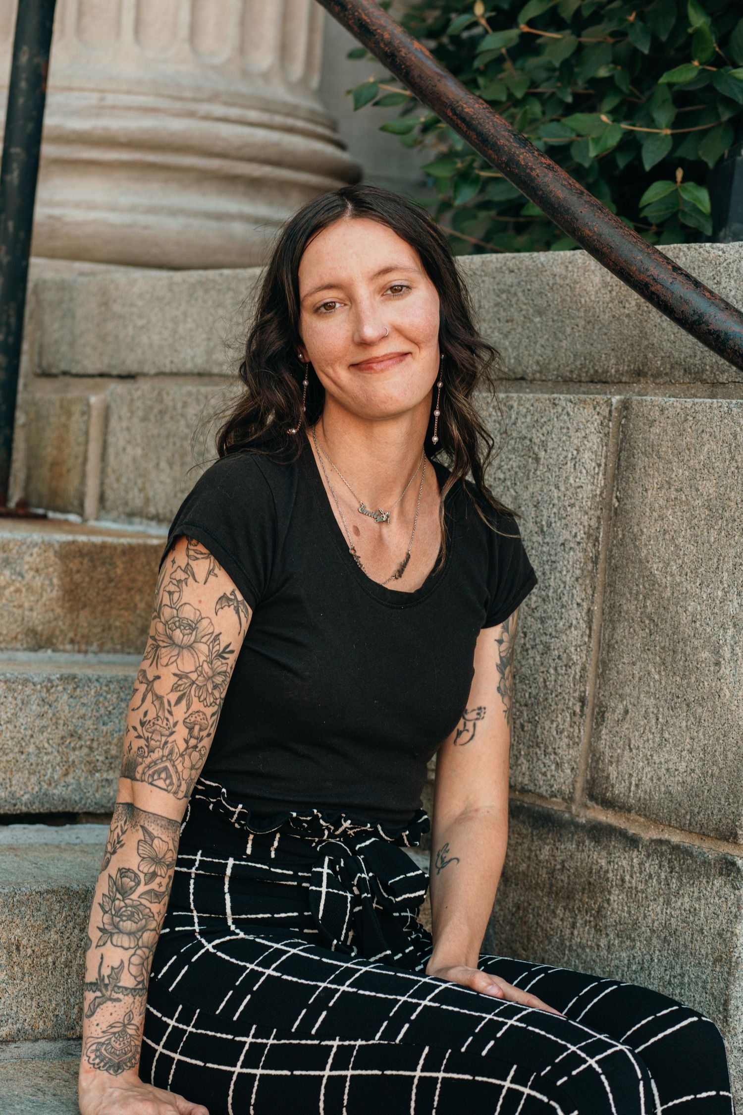 Tattooed woman with dark hair smiles while sitting on stone steps outdoors in black outfit.