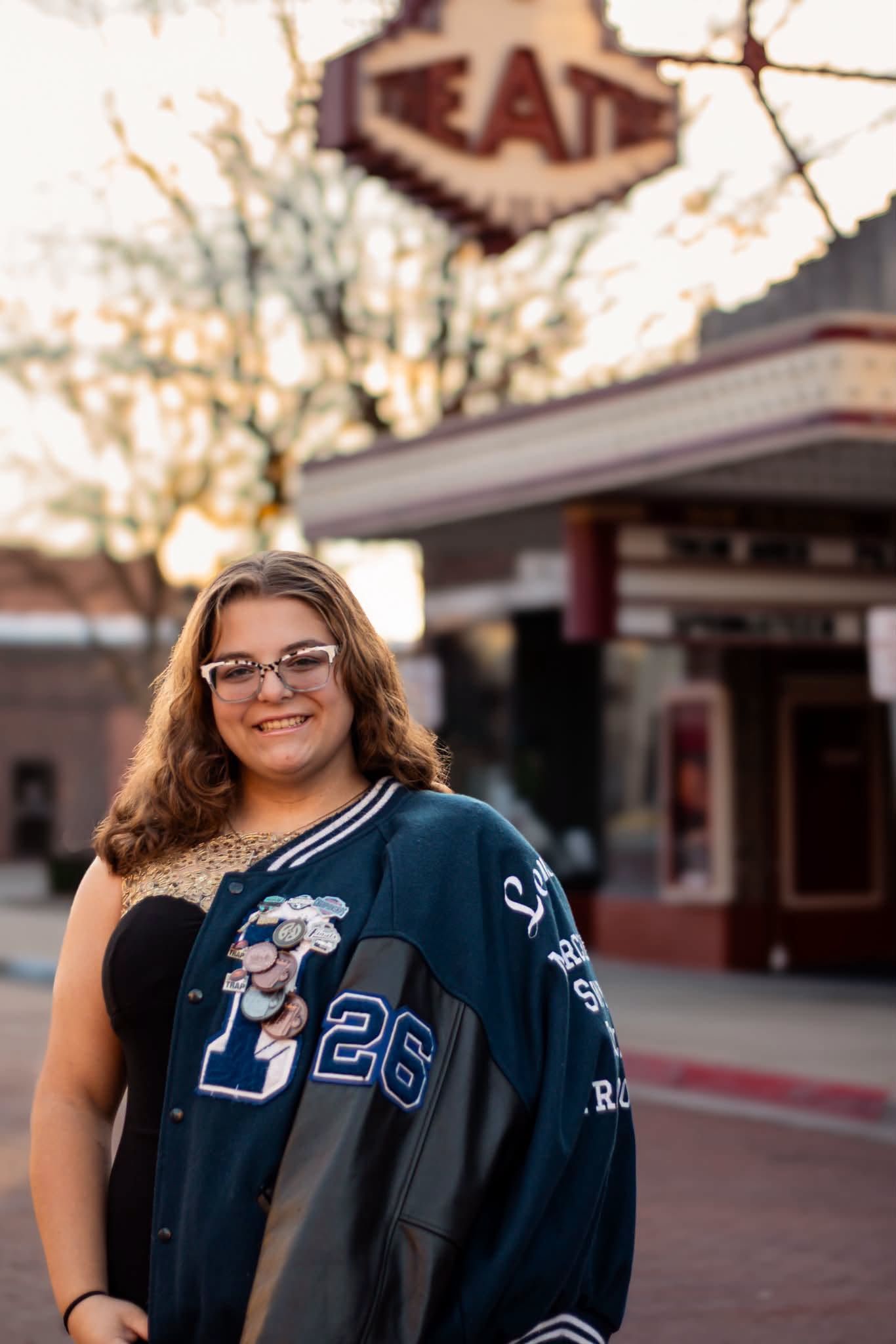 Teen girl in letterman jacket number 26 smiling outdoors near vintage theater sign at golden hour.