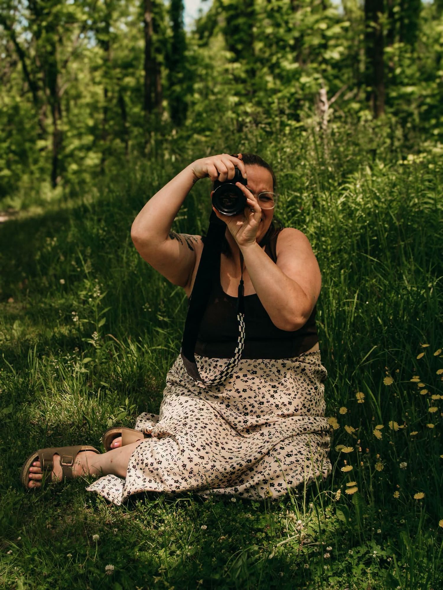 Woman sitting on grass in a forest, holding a camera up to her face, wearing a black top and floral skirt.