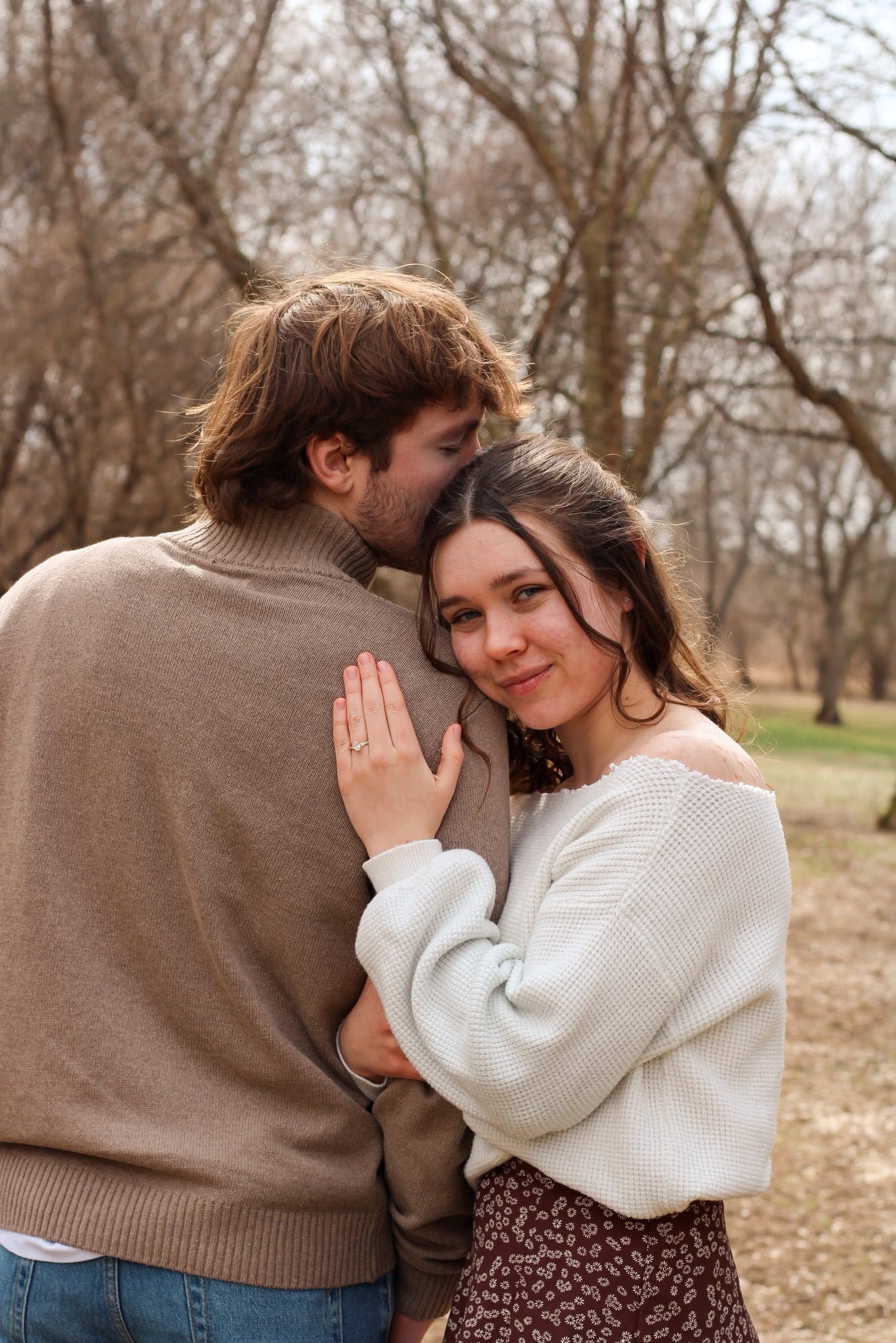 Happy couple embraces in a bare winter park, woman smiling at camera displaying engagement ring.