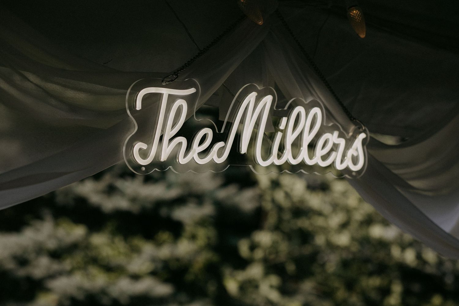 Glowing neon sign reading The Millers displayed under draped fabric ceiling at a wedding reception.
