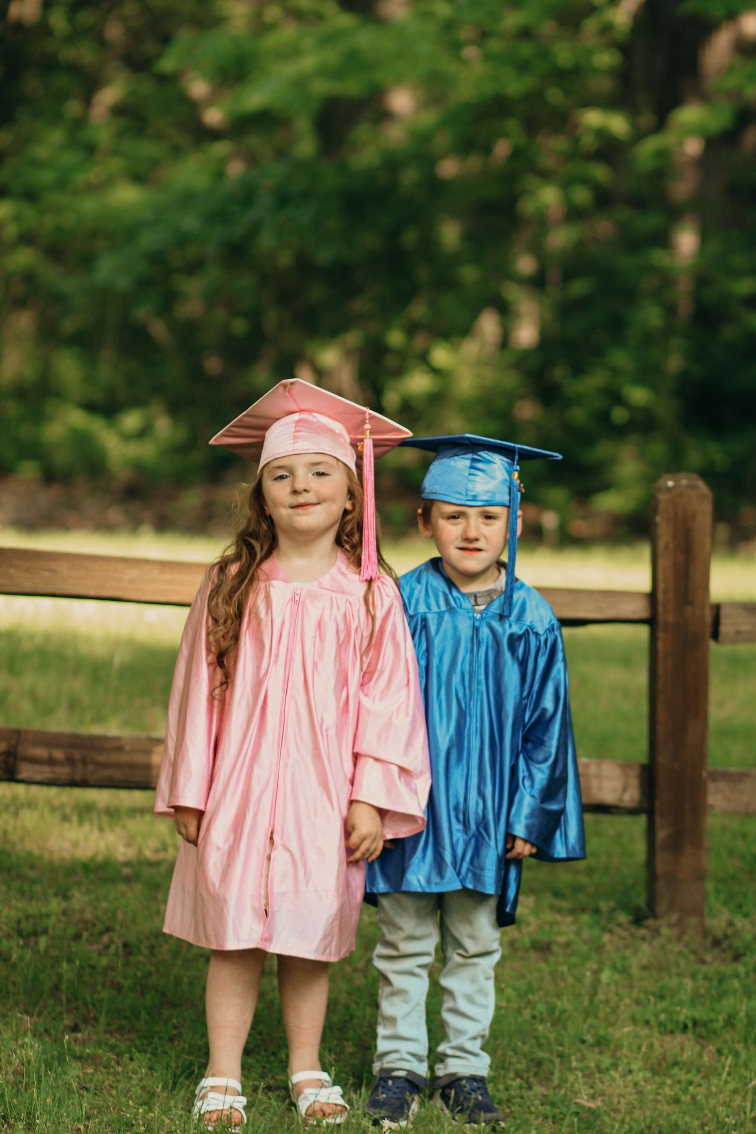 Two young children in pink and blue graduation caps and gowns posing outdoors near a wooden fence.