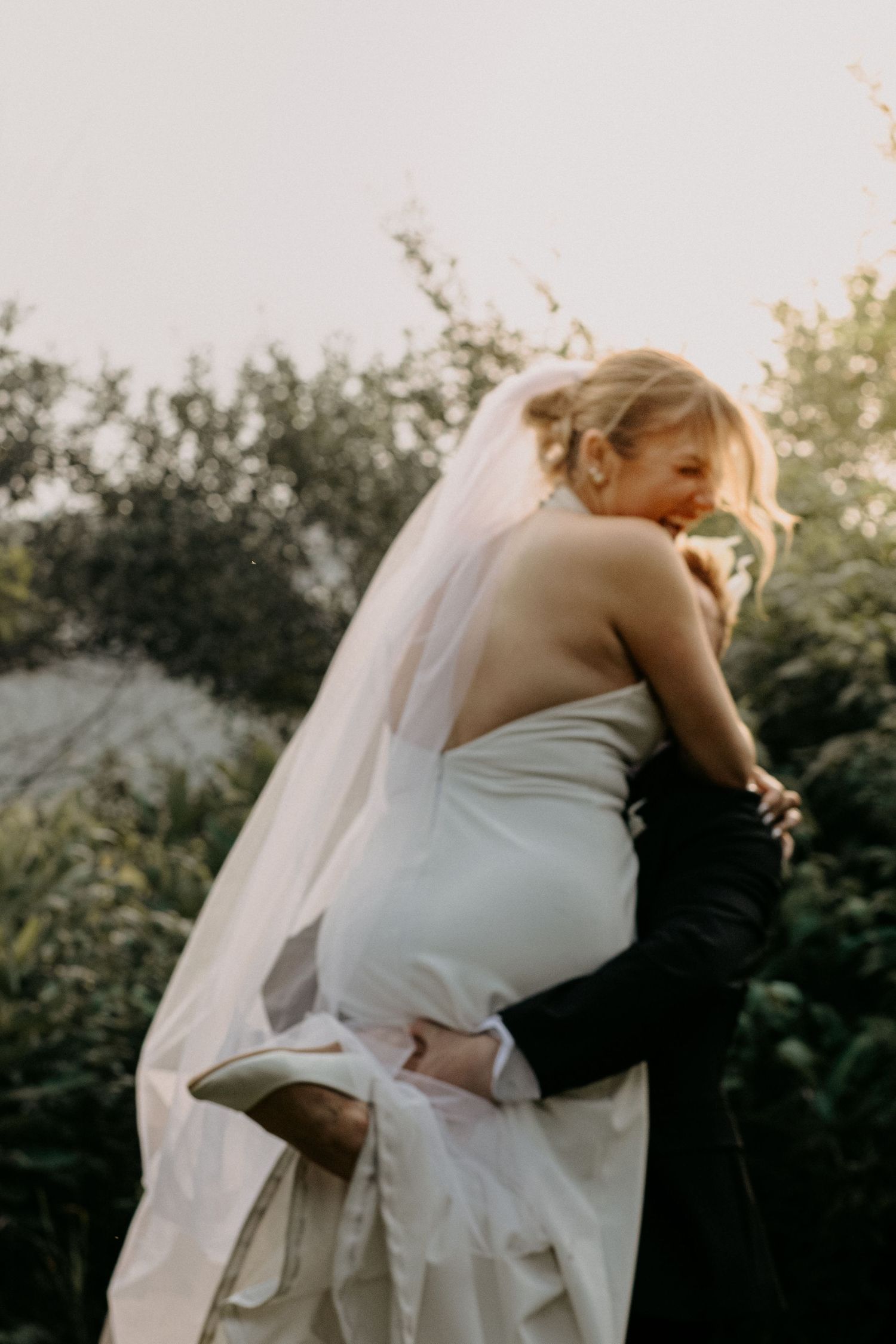 Bride in white gown and veil embracing groom outdoors at golden hour wedding ceremony.