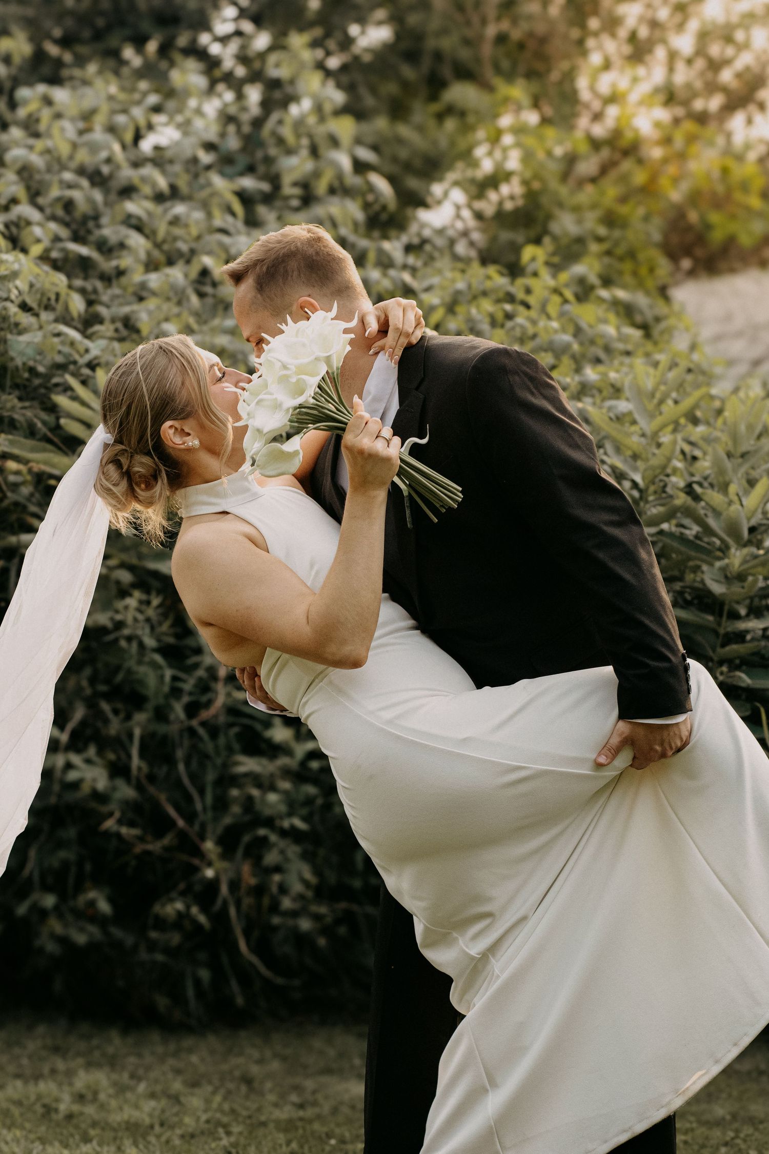Groom lifting bride in white halter gown with white bouquet for a romantic outdoor wedding portrait.