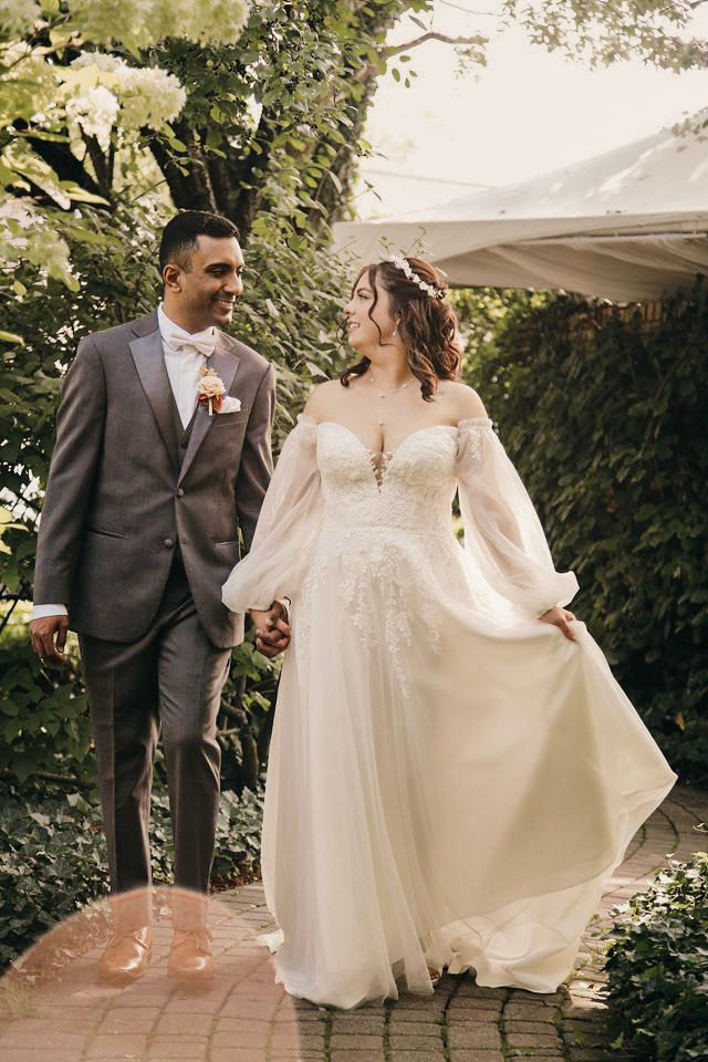 A bride and groom walk hand-in-hand through a lush garden, smiling at each other on their wedding day.