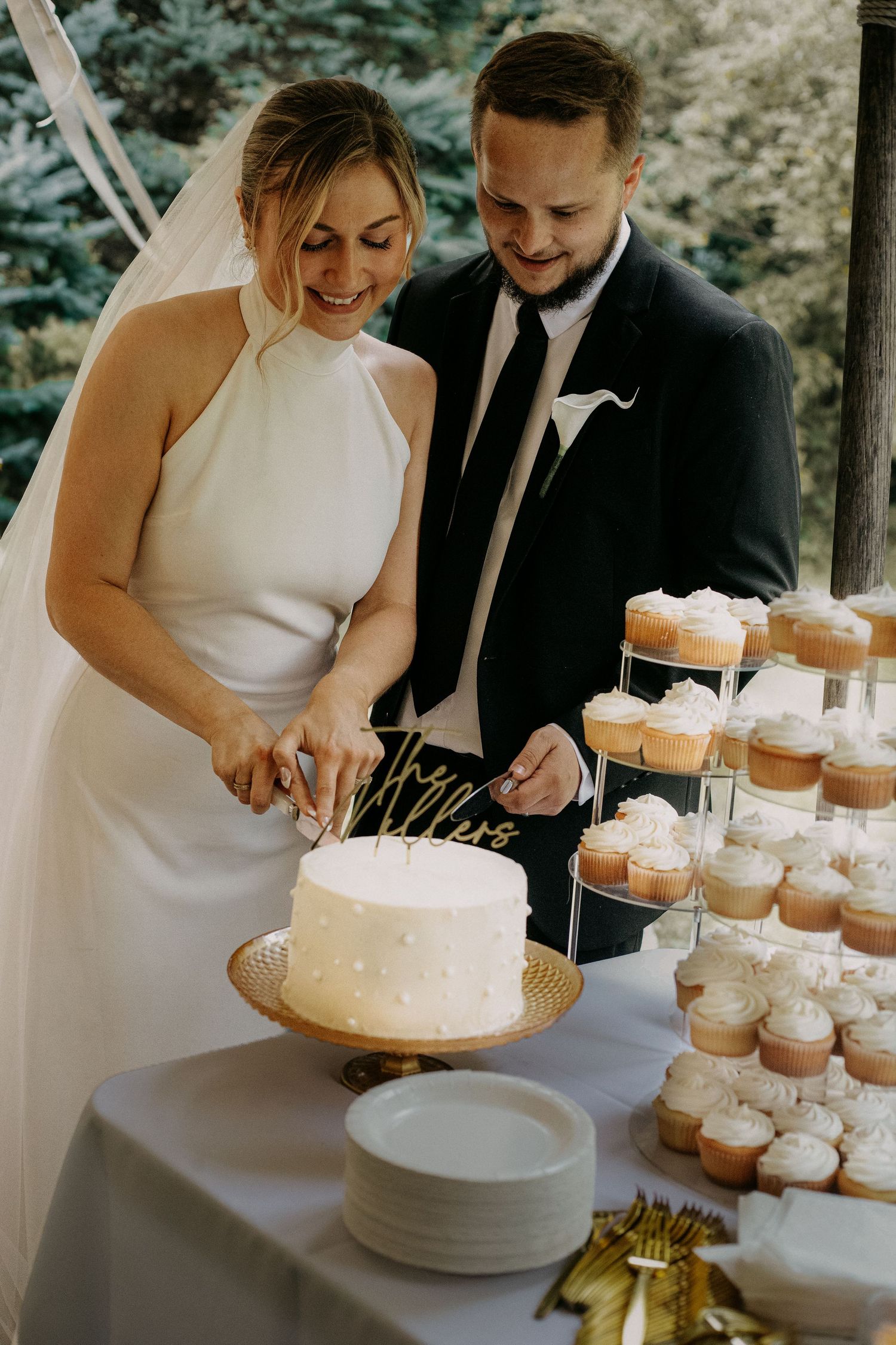 Bride and groom cutting white wedding cake at reception table with cupcake tower display.
