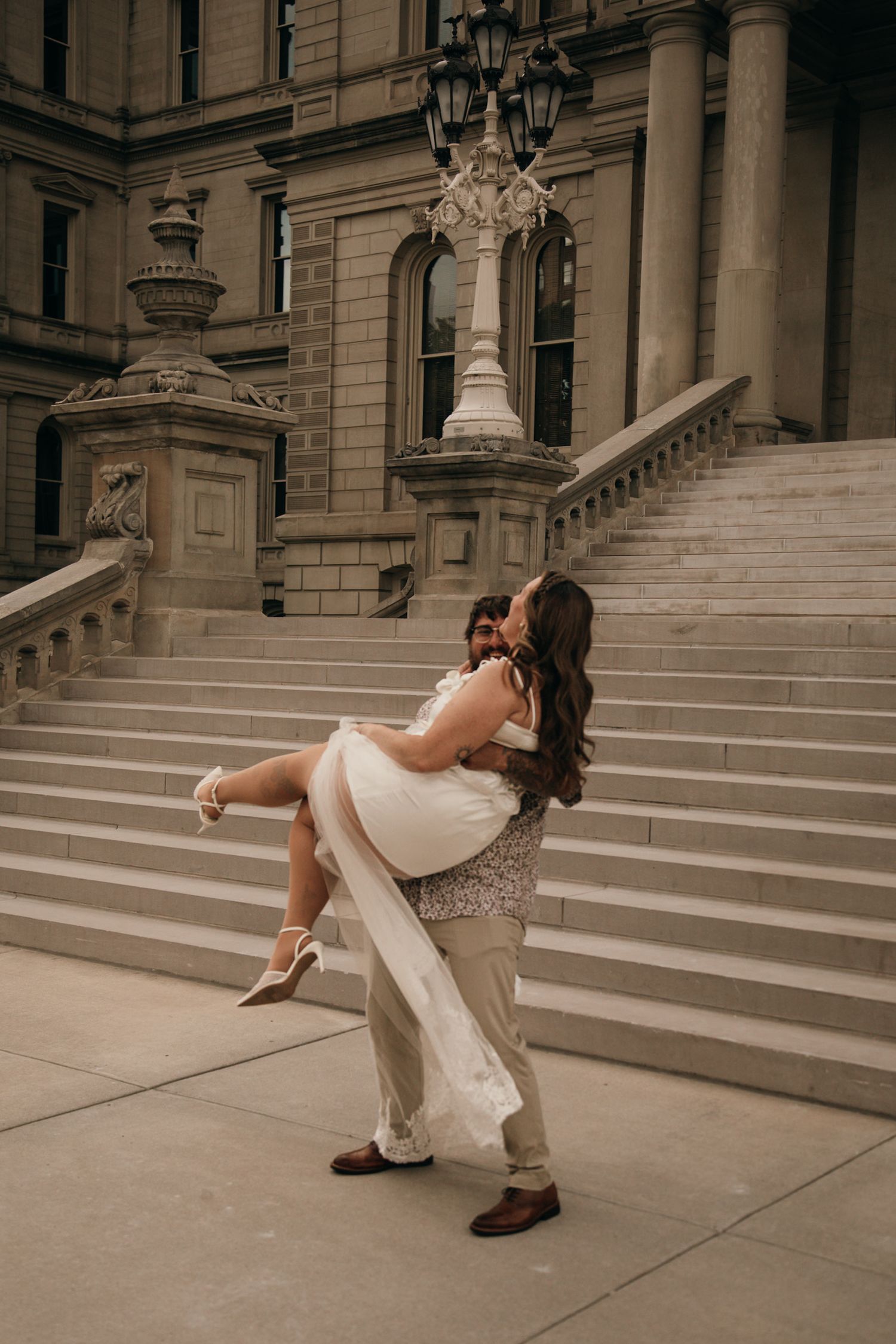 Groom lifting bride in white dress on grand stone steps during romantic outdoor wedding photo session.