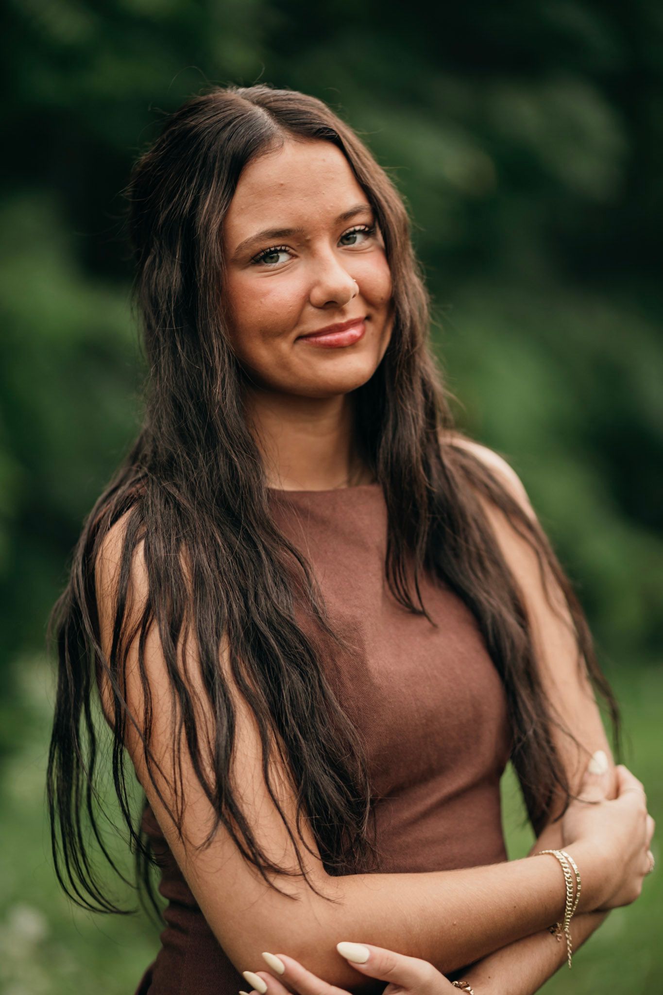Young woman with long dark hair in brown top smiling outdoors with green foliage background.