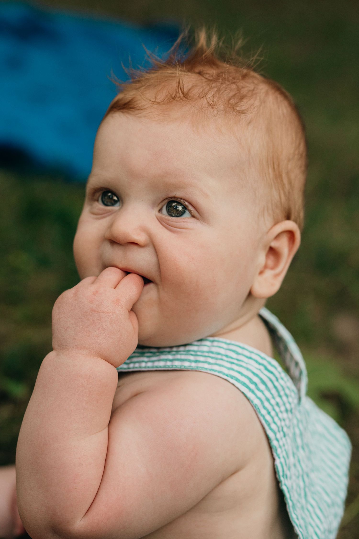 Adorable red-haired baby with fingers in mouth wearing striped outfit outdoors on grass.