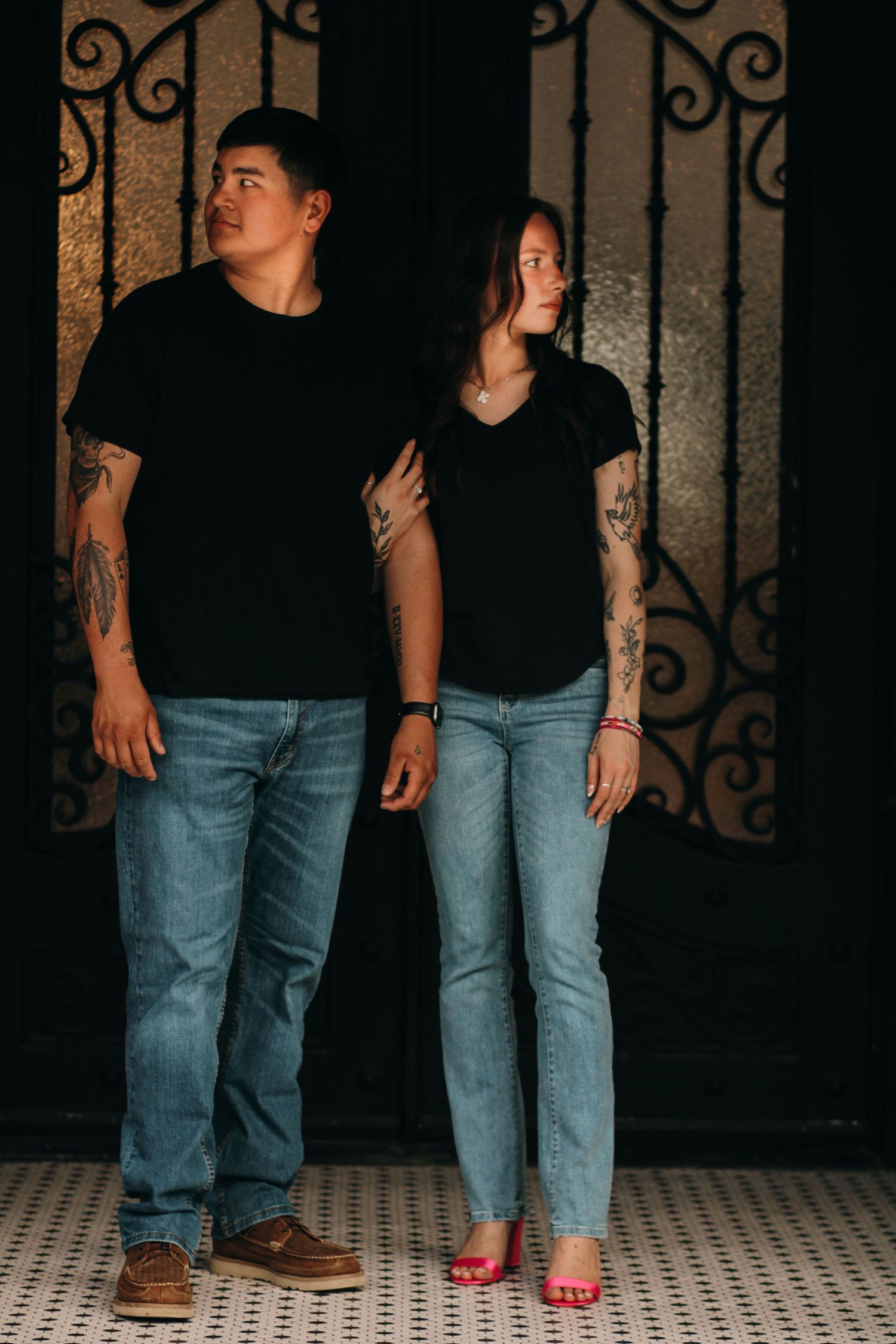 Couple in matching black shirts and jeans with tattoos standing by ornate iron door.