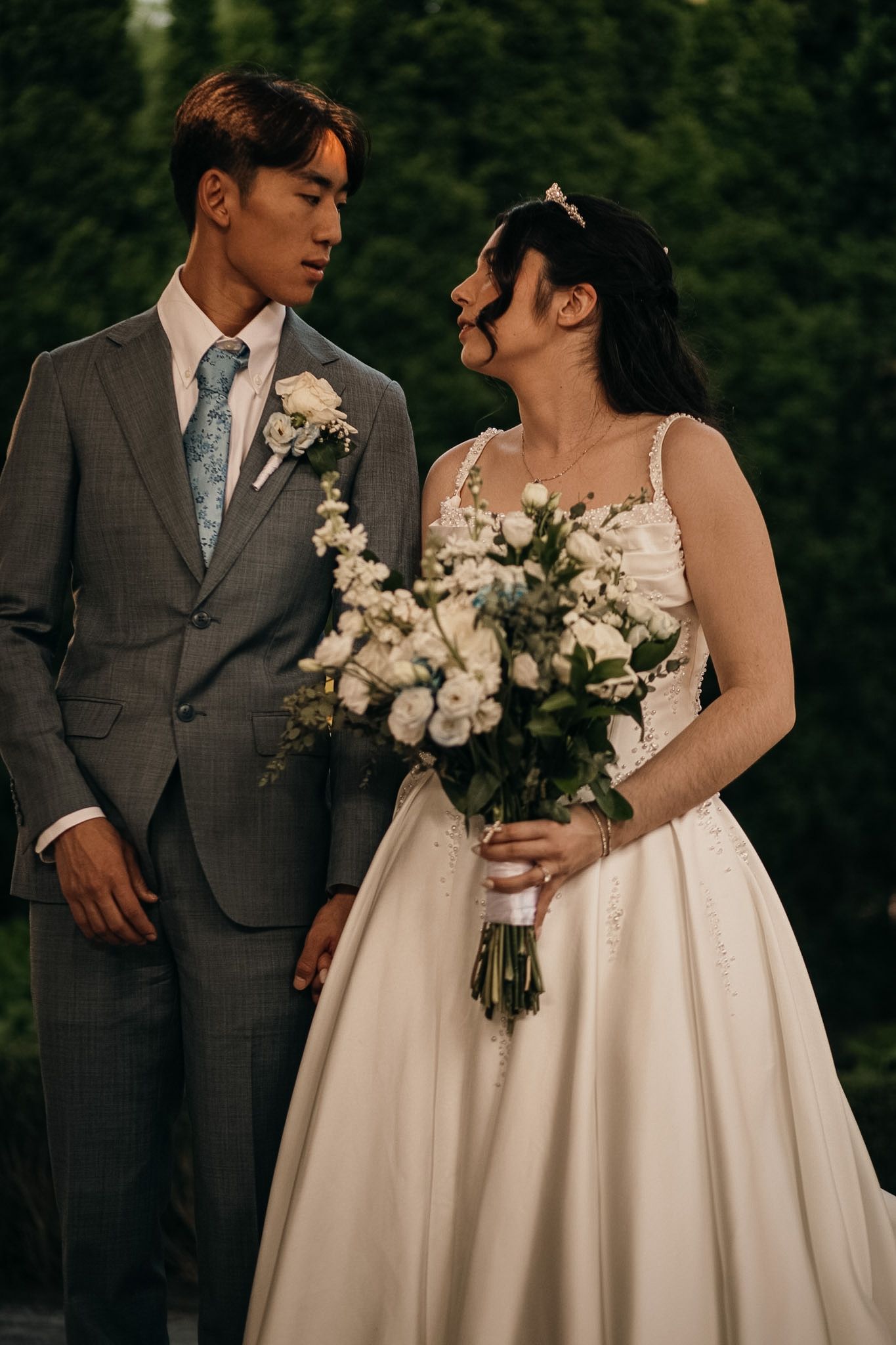 A bride in a white gown holds a bouquet of white flowers while a groom in a gray suit looks at her outdoors.