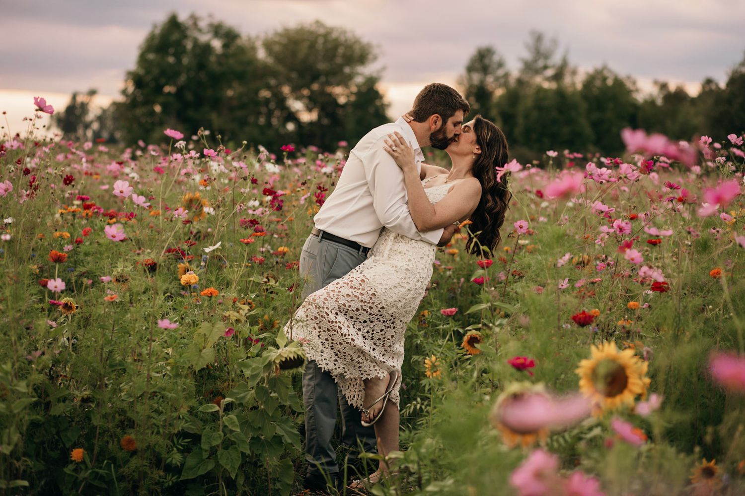 Couple sharing a romantic kiss in a vibrant flower field with pink blooms and sunflowers at dusk.