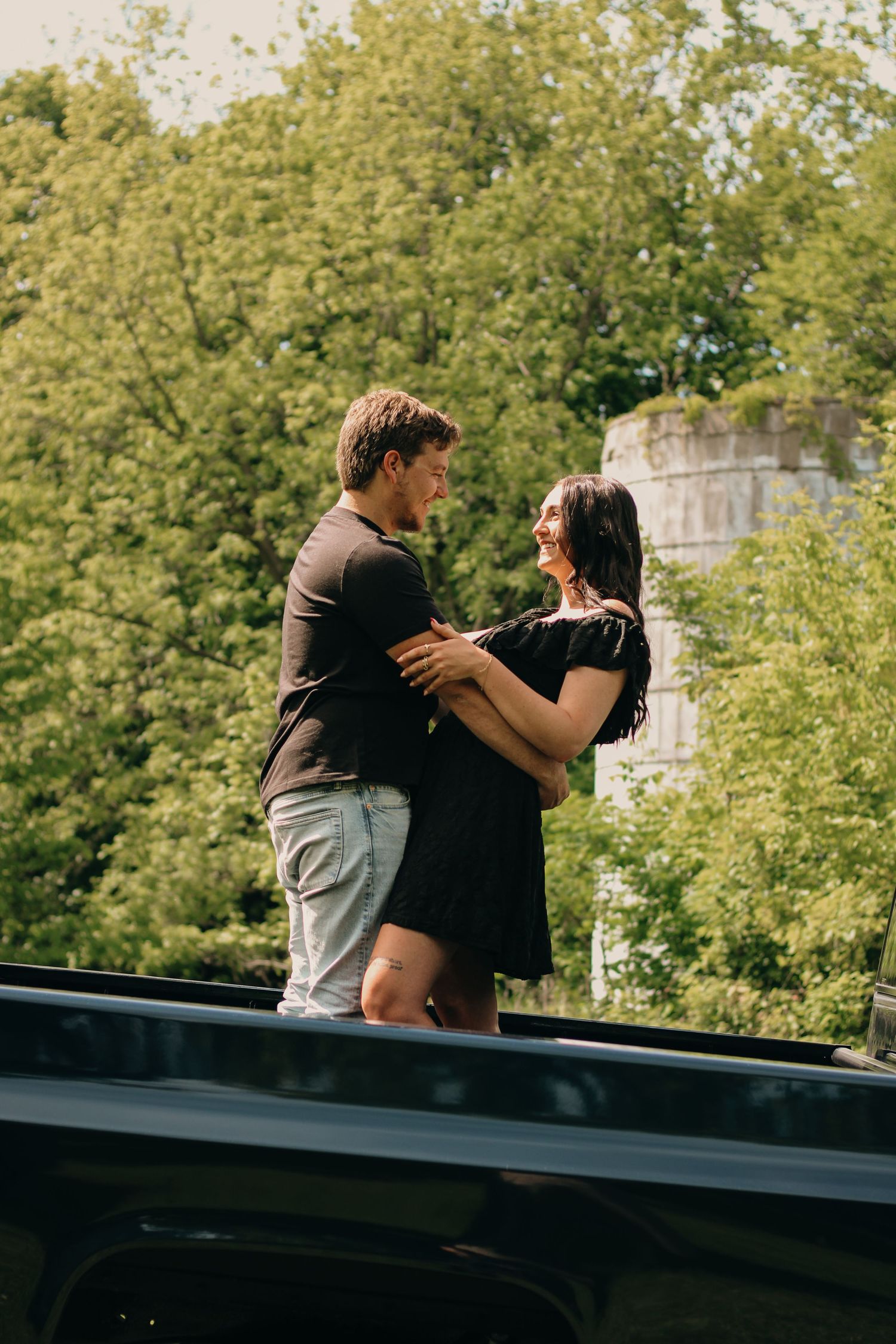 Young couple embracing and smiling at each other while standing in a truck bed with green trees behind.