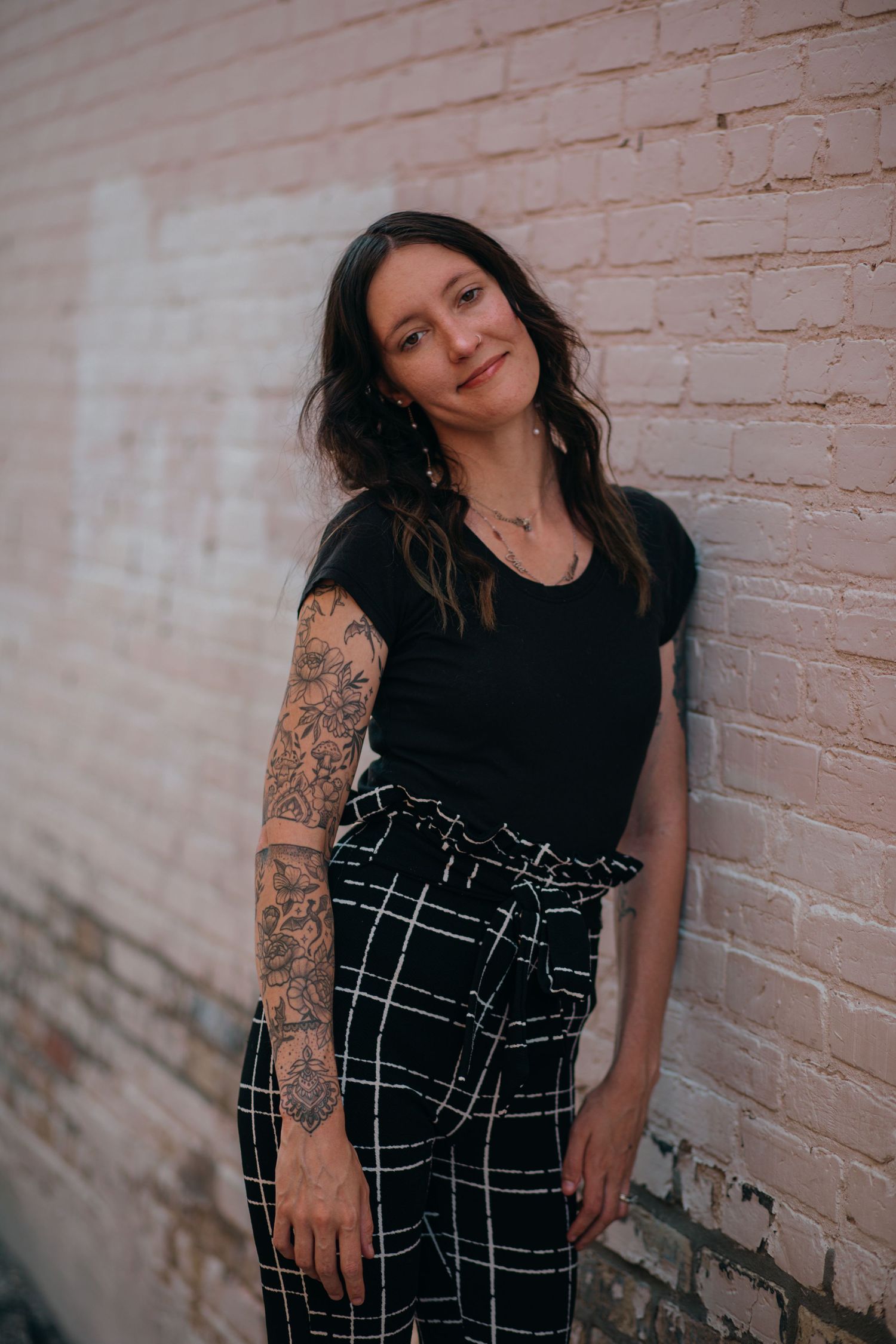Young woman with sleeve tattoos posing against a pink brick wall in black top and plaid pants.