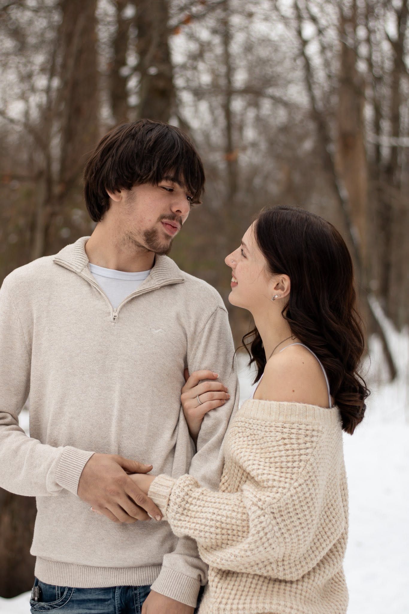 A couple in beige sweaters share a romantic moment in a snowy winter forest, gazing into each other's eyes.