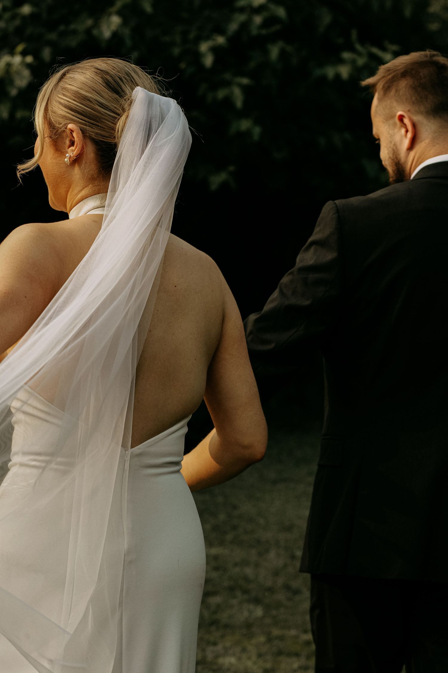 Bride in backless white halter gown with veil walking beside groom in black suit in dark garden.