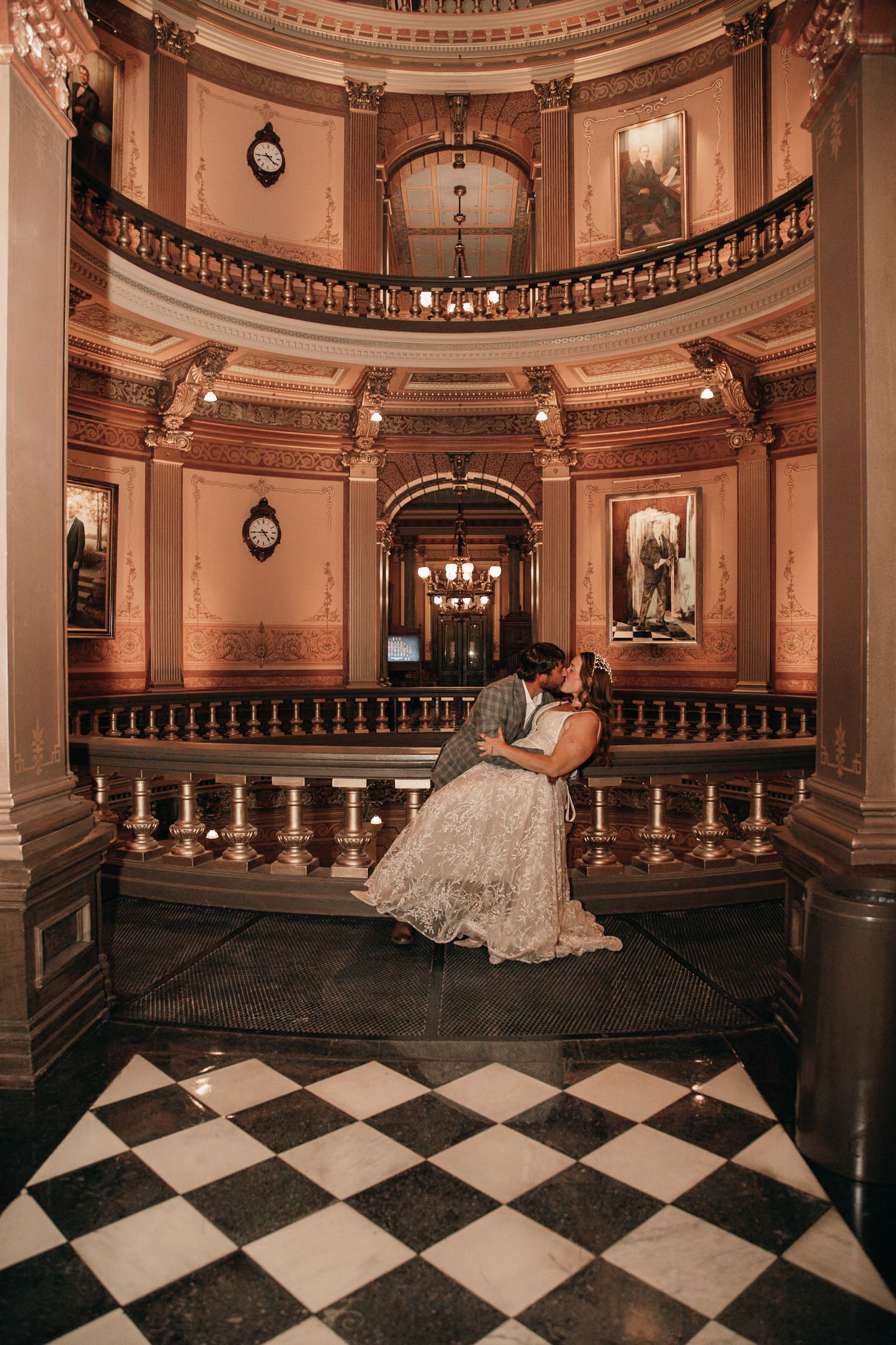 A couple shares a romantic kiss in an ornate grand hall with checkered floors and balconies.