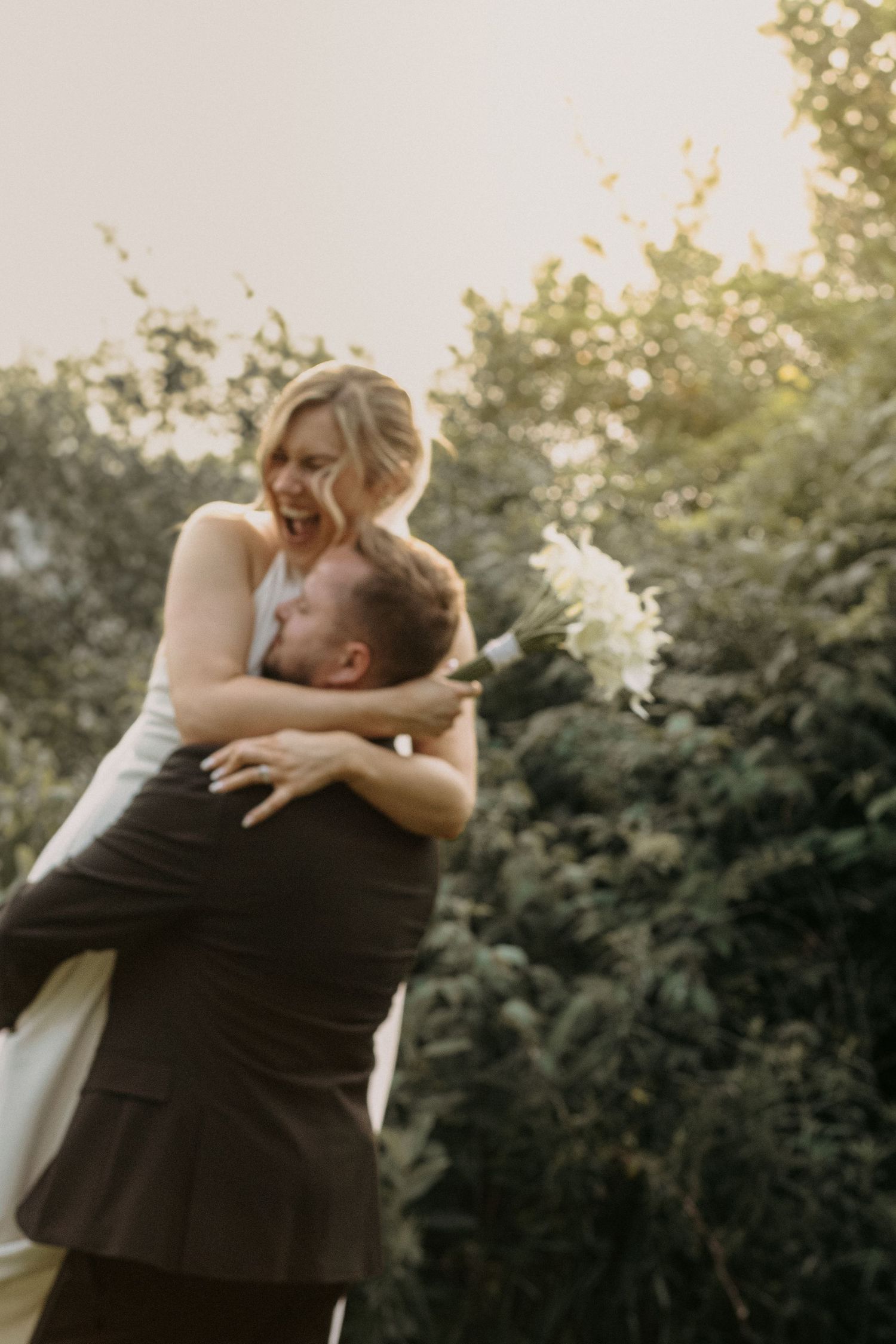 Joyful bride in white dress laughs as groom lifts her while holding white bouquet outdoors.