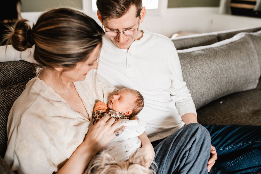 New parents sitting on a couch lovingly cradling their newborn baby together at home.