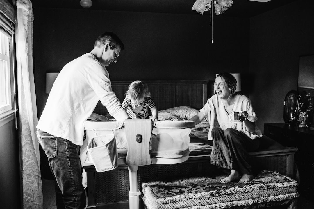 Family sharing a joyful moment in a bedroom, with a child between parents on a bed in black and white.
