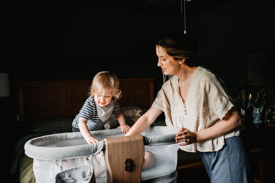 Young mother and toddler child looking into a modern white bassinet in a dark-walled bedroom.