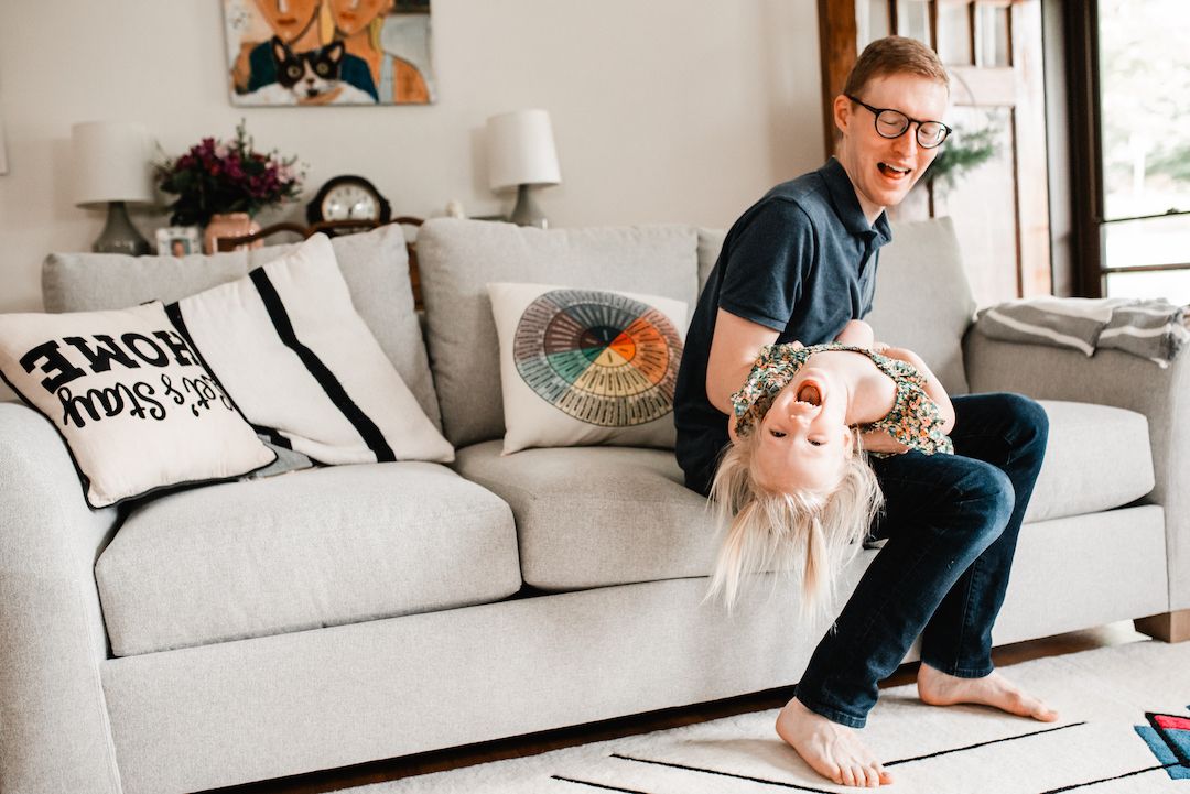 A father playfully flips his laughing child upside down on a couch in a bright living room.