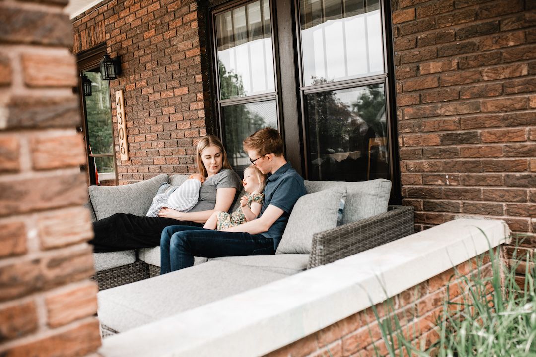 Family of three reading together on an outdoor couch on a brick home porch.