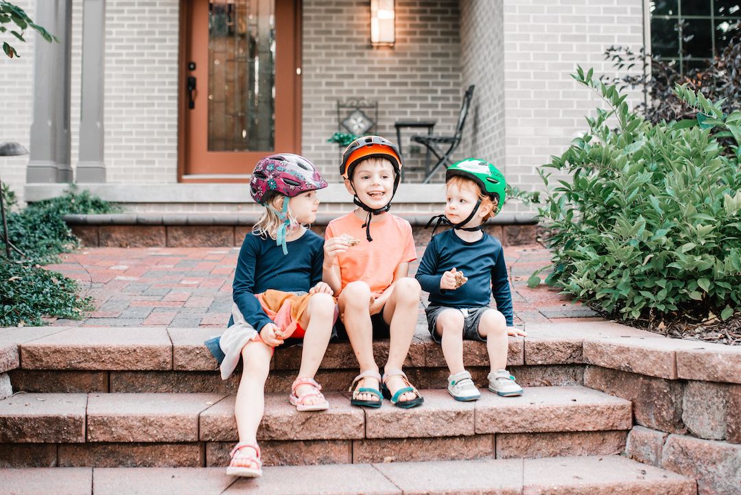 Three children in colorful bike helmets sitting together on front porch steps.