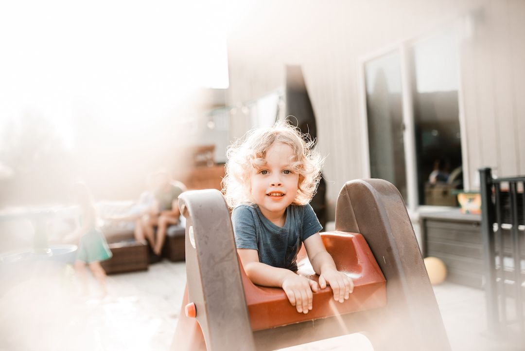 Curly-haired toddler playing on indoor slide in bright sunlit living room.
