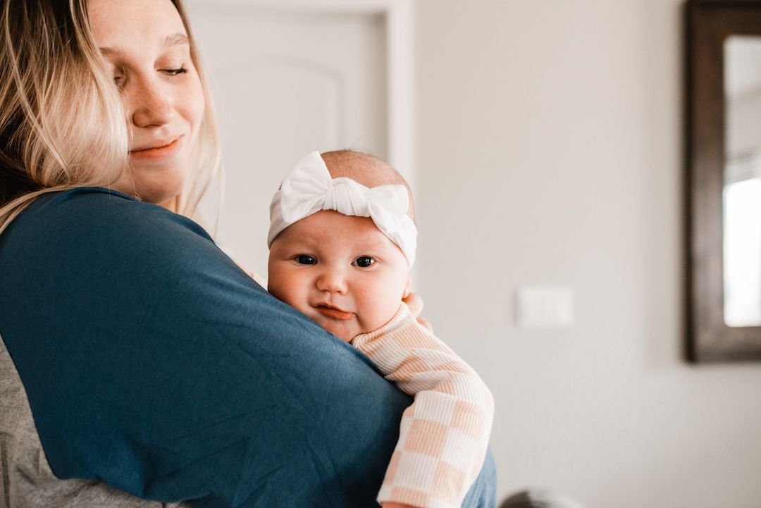 Mother tenderly holds smiling baby girl wearing white bow headband against her shoulder indoors.