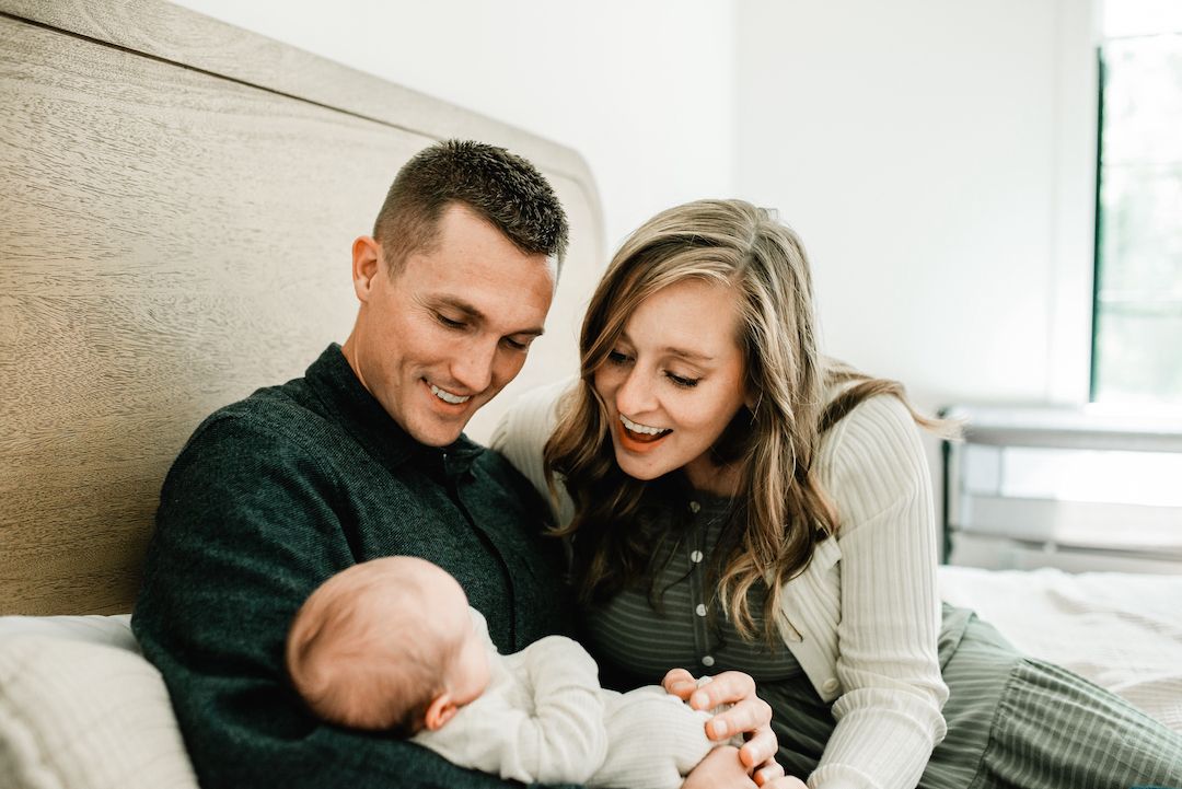 Smiling couple sits on a bed, joyfully looking down at their newborn baby in a bright bedroom.