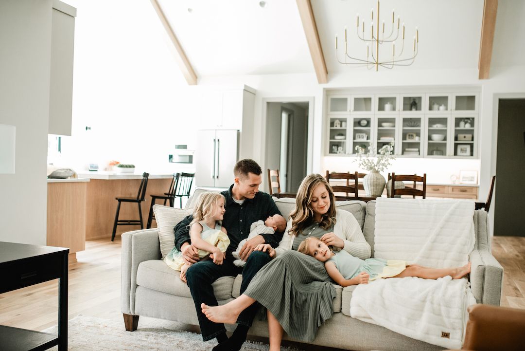 Family of four with newborn twins sitting on a cozy gray sofa in a modern open-plan living room.