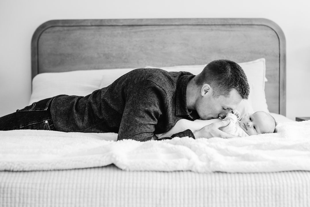 Black and white photo of a father lying on a bed, gently kissing his newborn baby wrapped in a white blanket.