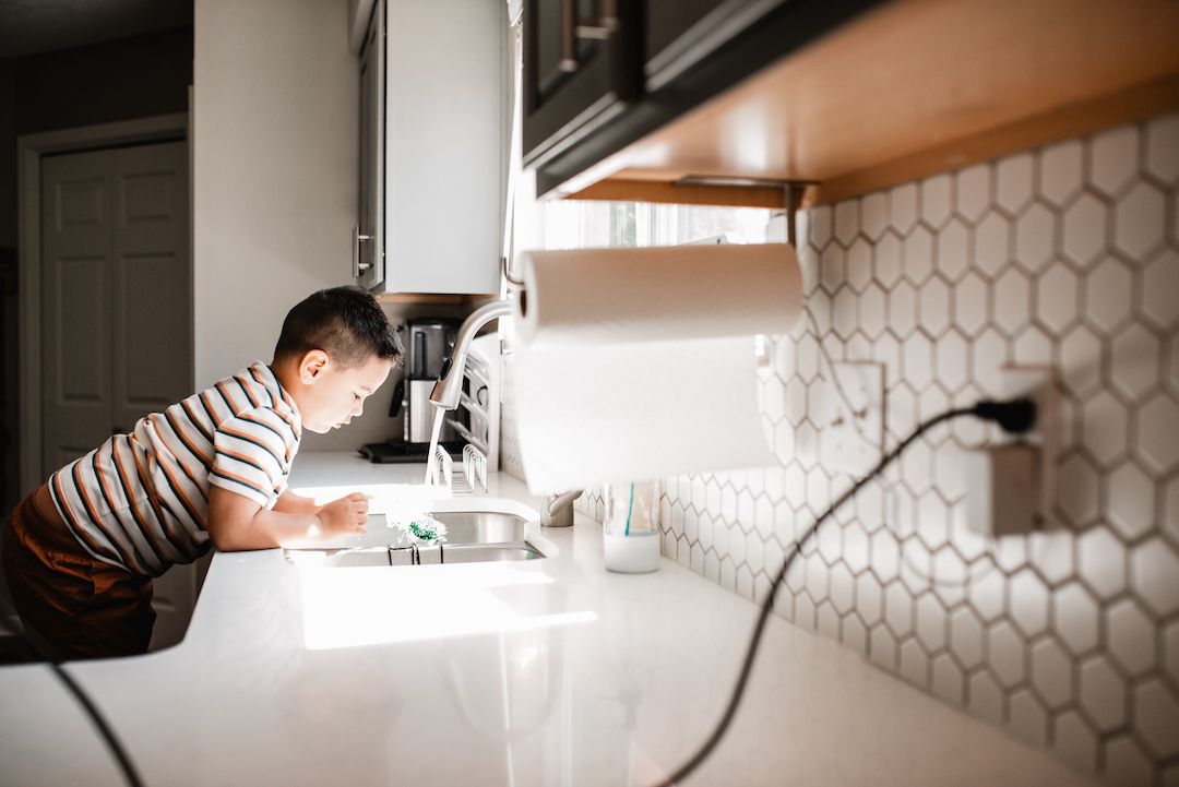 Young boy leaning over kitchen sink washing hands in modern white kitchen with hexagon tile backsplash.
