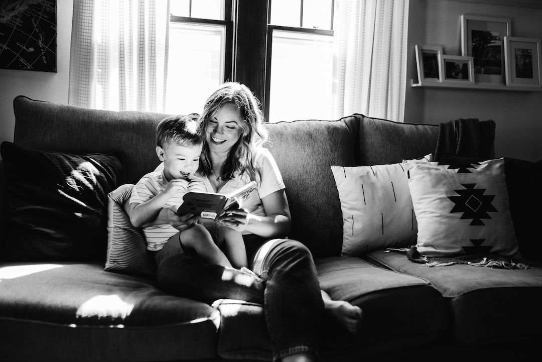 Black and white photo of a mother and young son reading a book together on a cozy living room couch.