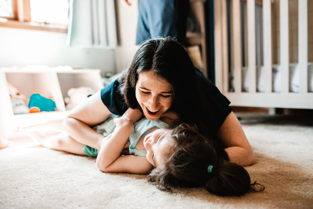 A laughing mother playfully lies on top of her young daughter on a cozy carpeted floor near a white crib.