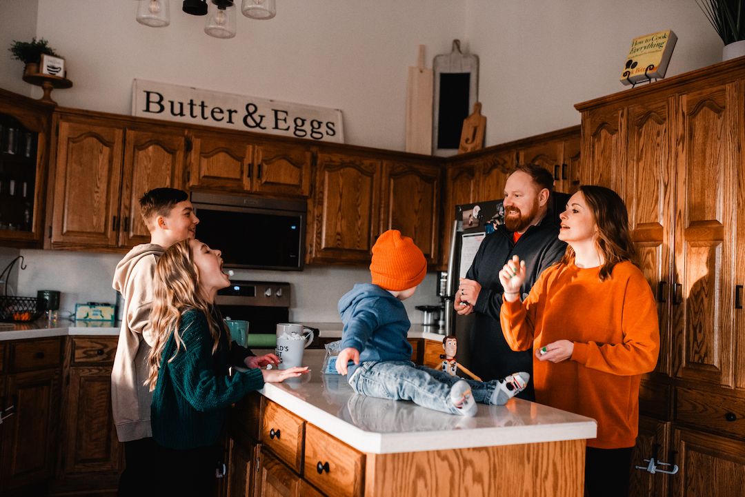 Family gathering in a rustic kitchen with a Butter and Eggs sign, parents and kids sharing a fun moment.