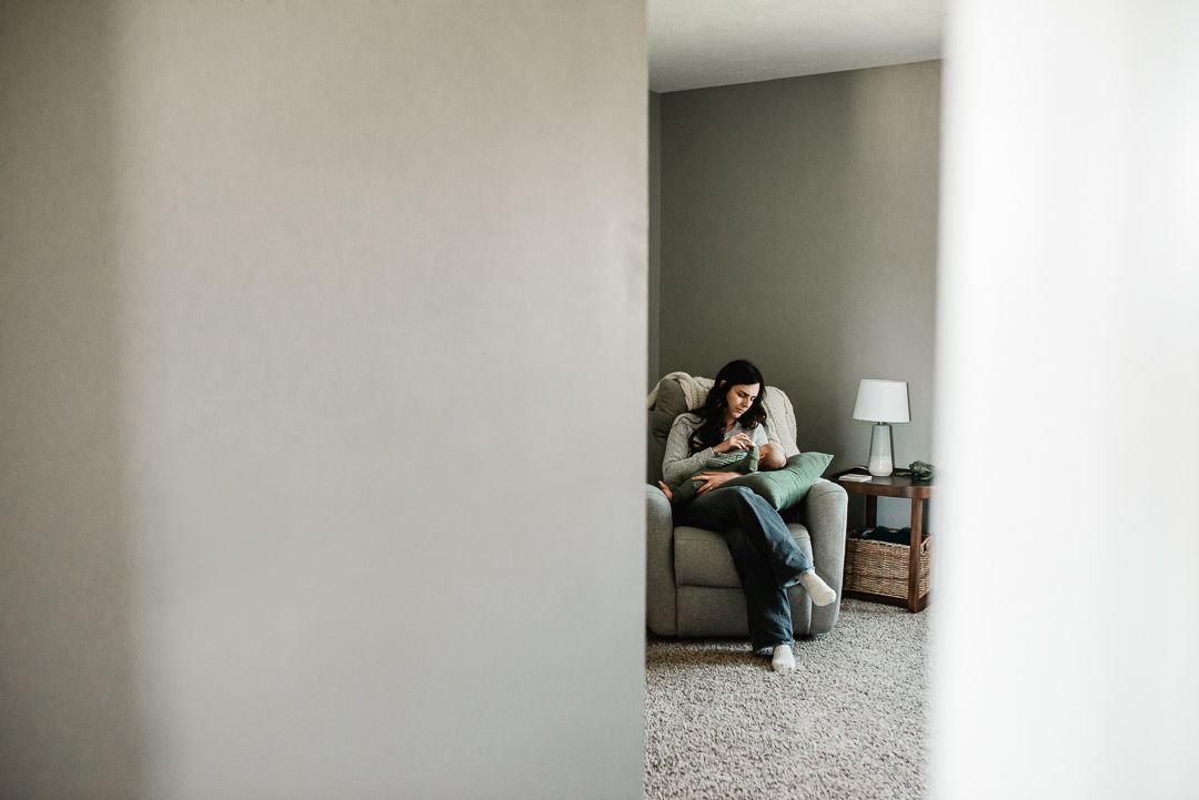 Woman sitting alone on a cozy armchair in a softly lit living room, viewed through a doorway.