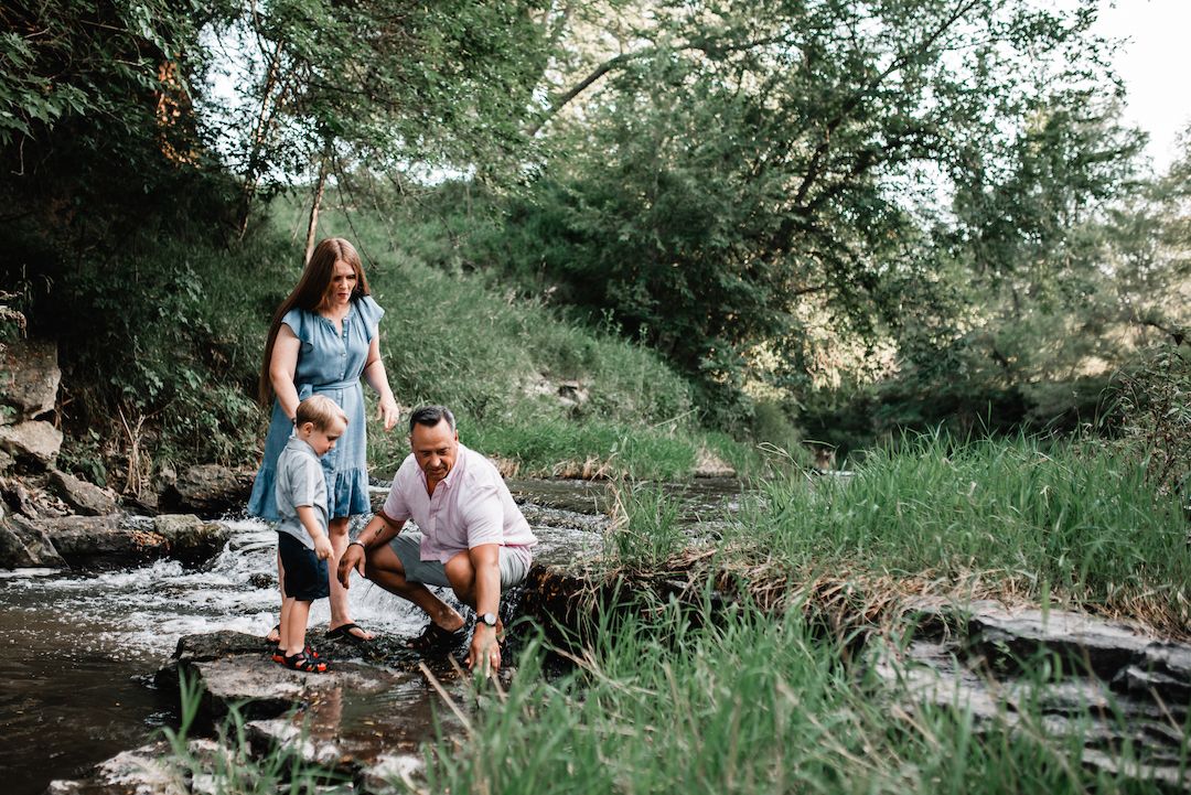 Family of three exploring a rocky creek surrounded by lush green trees in summer.