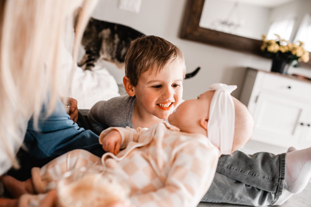 Happy young boy smiling while holding a newborn baby in a cozy home setting with family nearby.