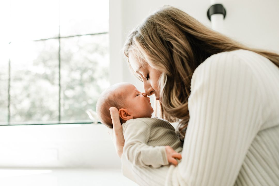 A tender moment as a young mother holds her newborn baby close, touching noses in a bright, airy room.