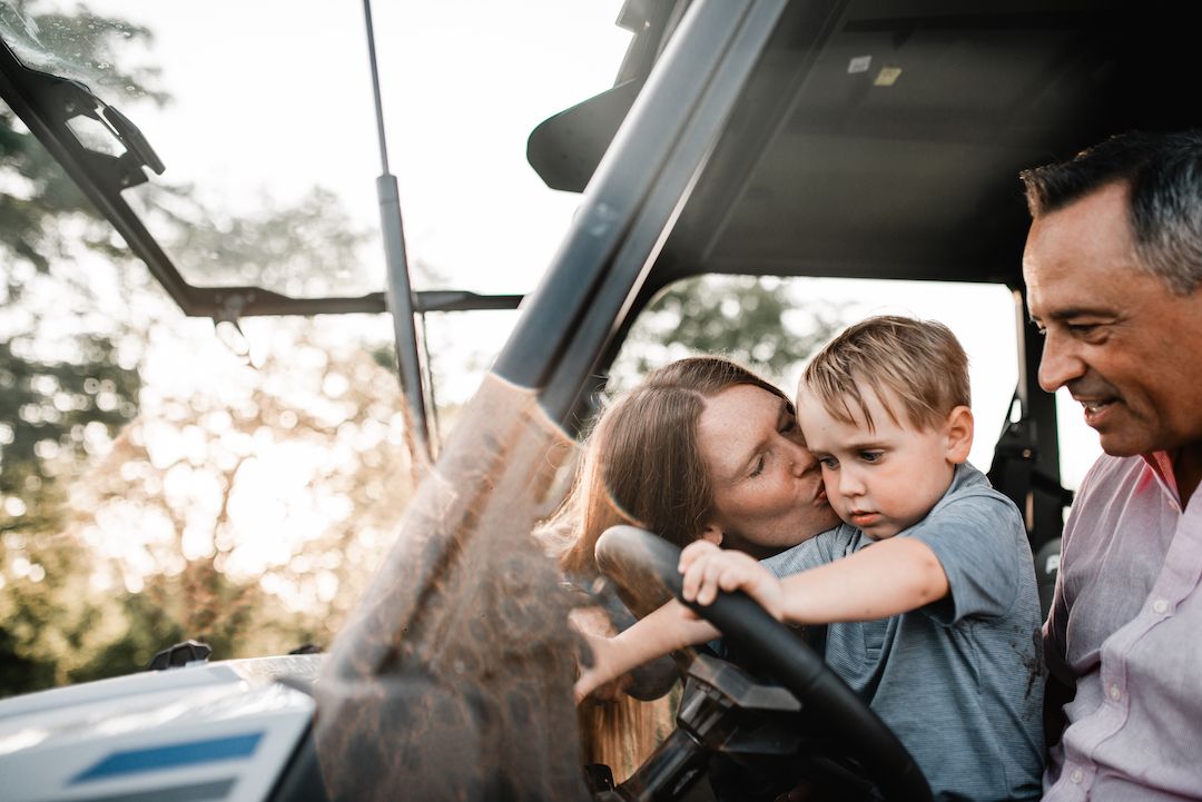 Mother kissing young boy on the cheek while sitting in an open vehicle with grandfather smiling.