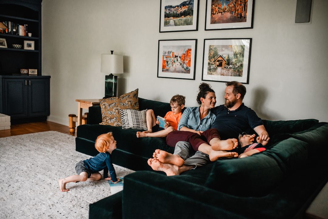 Family of four relaxing together on a dark green velvet sofa in a cozy living room.