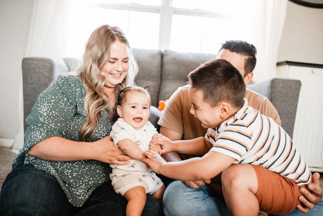 Happy family sitting on floor with mom, dad, older child, and laughing baby.