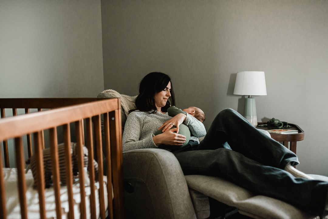 Mother holds newborn baby while relaxing in gray nursery glider chair.