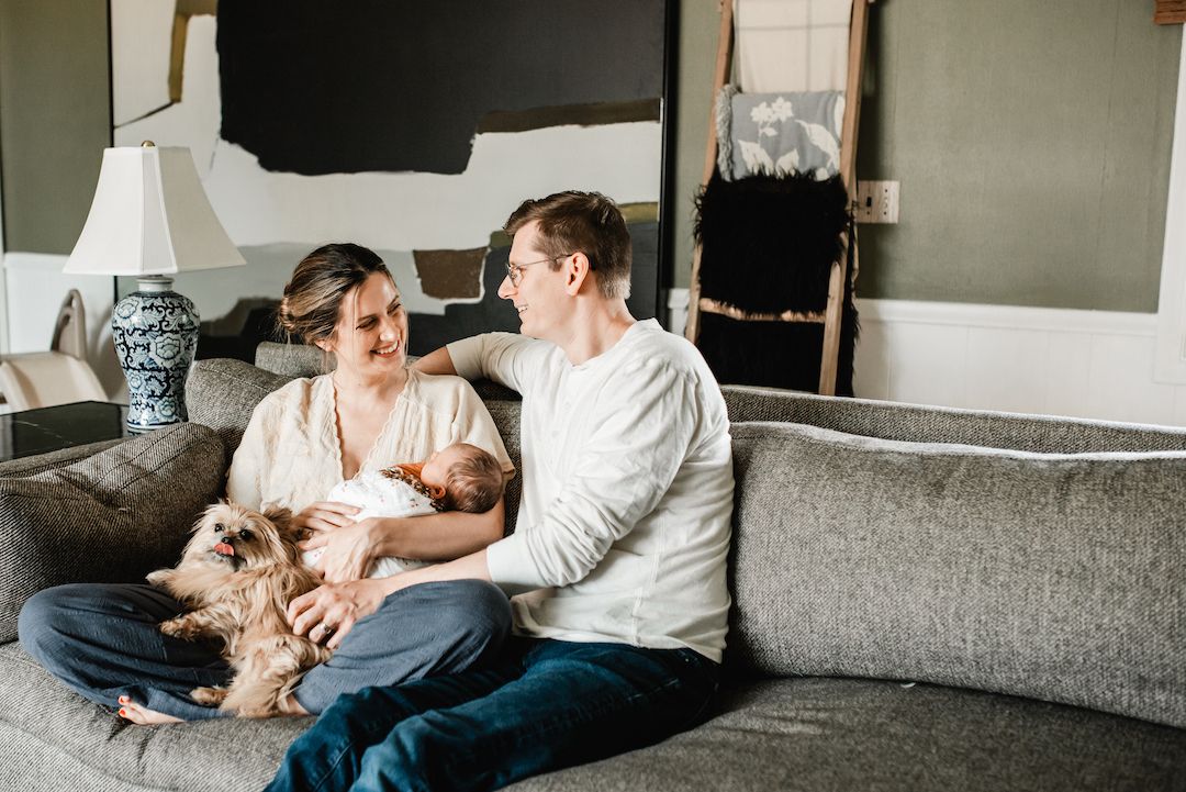 Couple sitting on gray sofa with newborn baby and small dog in cozy living room.